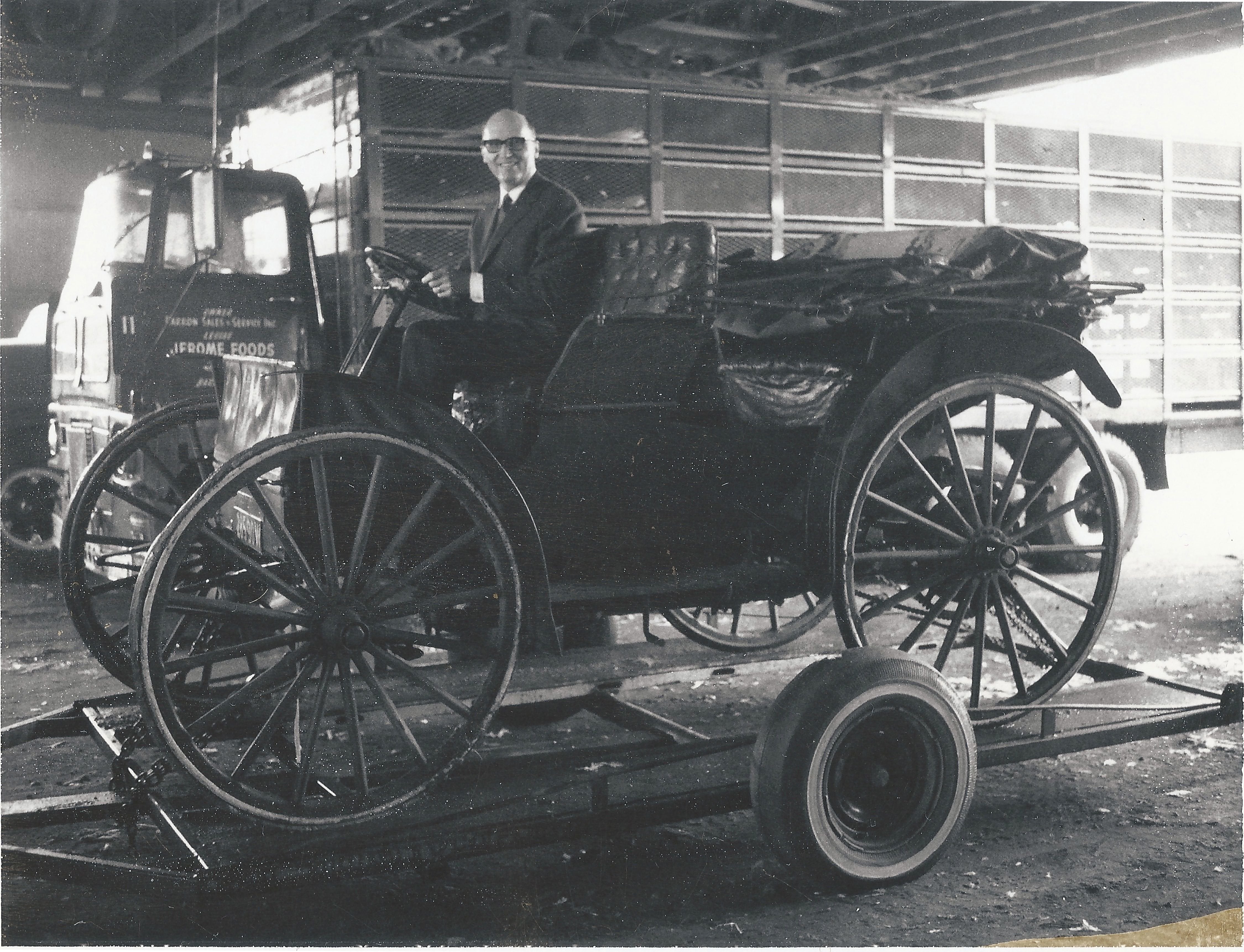 Black and white photo of a man in a suit smiling in an antique car on a trailer, with a truck behind him.