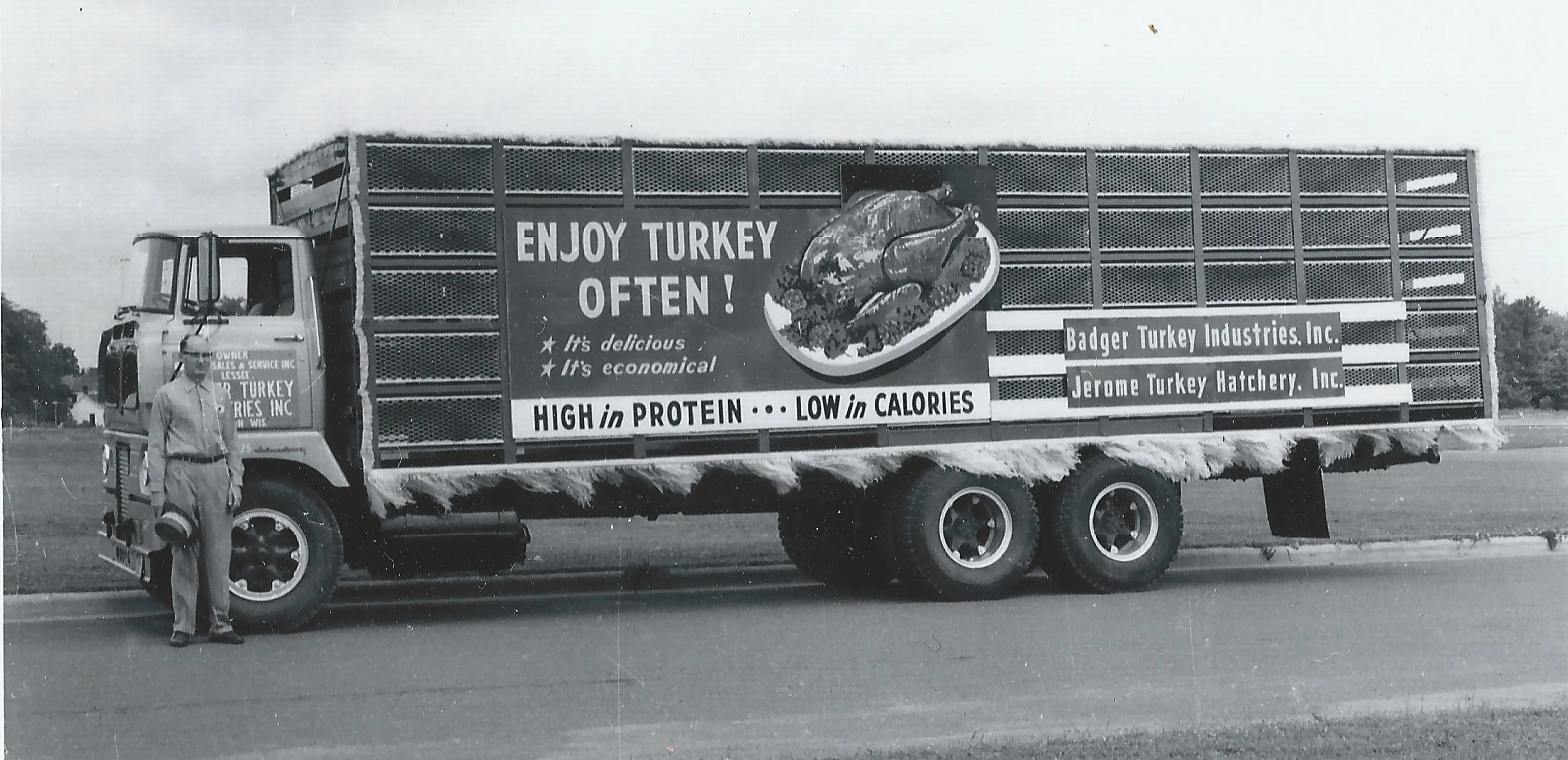 Black and white photo of a man next to a vintage truck with a promotional sign for turkey on its mesh-sided cargo bay.
