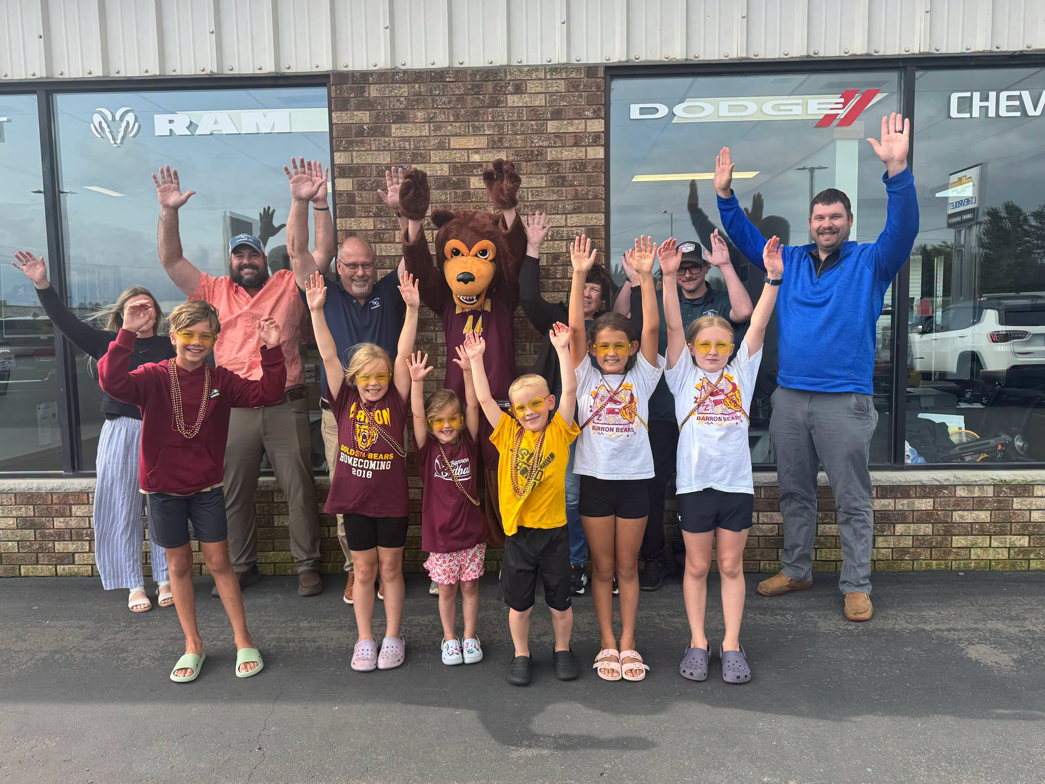 Group of smiling adults, children, and a bear mascot raise their hands in front of a car dealership.
